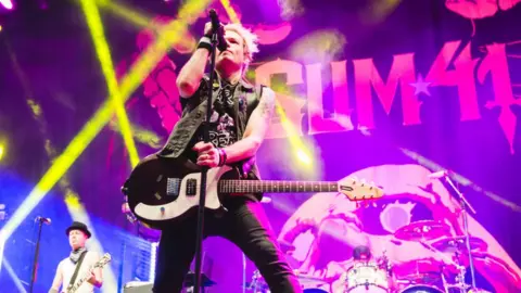 Getty Images Deryck Whibley of Sum 41 performs on stage during day 3 of Download festival 2019 at La Caja Magica on June 30, 2019 in Madrid, Spain. Deryck is a white man in his 40s with bleached blonde hair. He's wearing a sleeveless black denim jacket over a black top, paired with tight black jeans. He has a guitar slung over his shoulder and holds a microphone with his right hand. The staging is lit by pink and yellow lights with the band's logo behind Deryck.
