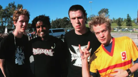 Getty Images Sum 41 pictured in 2001. The band at the time was made up of (L-R) Jason McCaslin, Dave Baksh, Steve Jocz and Deryck Whibley. The band are in their early 20s and are photographed outside in a car park on a blue-sky day. Jason is a white man with red hair wearing a black T-shirt. Dave is an Asian Canadian man with grown out black hair and a nose piercing and a black T-shirt. Steve is a white man with cropped dark hair, also wearing a black T-shirt. Deryck is a white man with spiky blonde hair, wearing a yellow T-shirt with a red stripe.