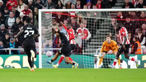 PA Media Iliman Ndiaye wears a black kit. He can be seen kicking a ball into a goal on a football pitch as a player in red and white jerseys tries to intercept. They are on a green football pitch and the goal keeper wears an orange jersey. 